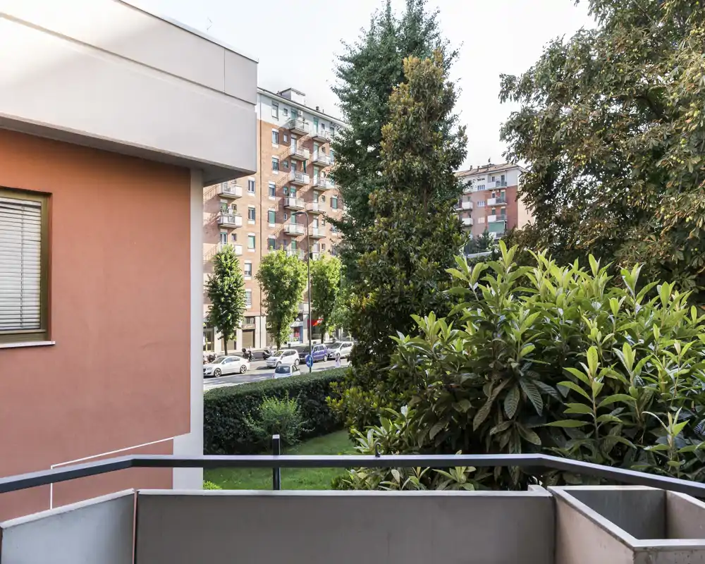 Sunny balcony view overlooking a tree-lined street and neighboring buildings, with a small railing and planted greenery in the foreground.