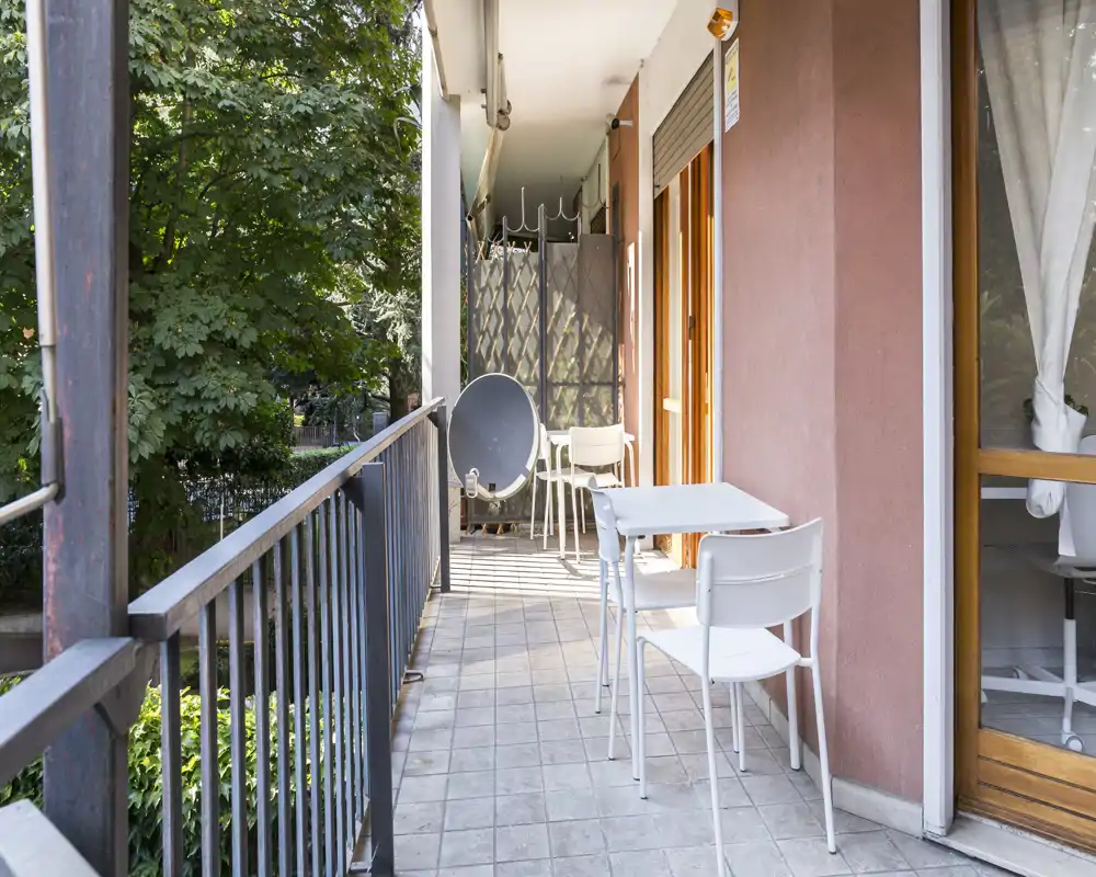 Sunny narrow balcony with a small table and two chairs, tiled floor and railing overlooking trees — suitable for morning coffee.