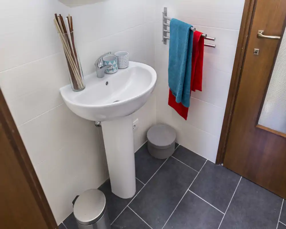 Small, modern bathroom with a pedestal sink, towel rail with colorful towels, dark tiled floor and white wall tiles; clean and well-lit.