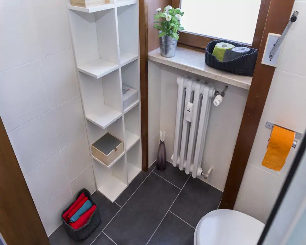 Small, well-kept bathroom corner with tiled floor, radiator under the window, shelving for toiletries and a toilet partially visible.