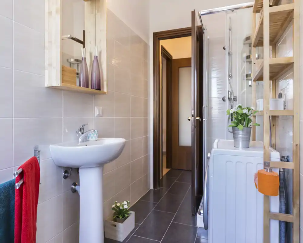 Modern, tidy bathroom with pedestal sink, shower cabin and washer; neutral tiles and wooden shelving create a clean, functional space.