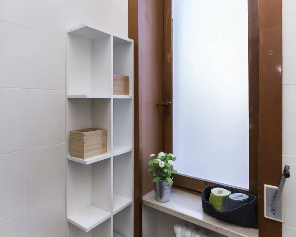 Compact bathroom corner with a frosted window, a white cubed shelving unit and a small plant on the sill; clean, bright and neatly organized.
