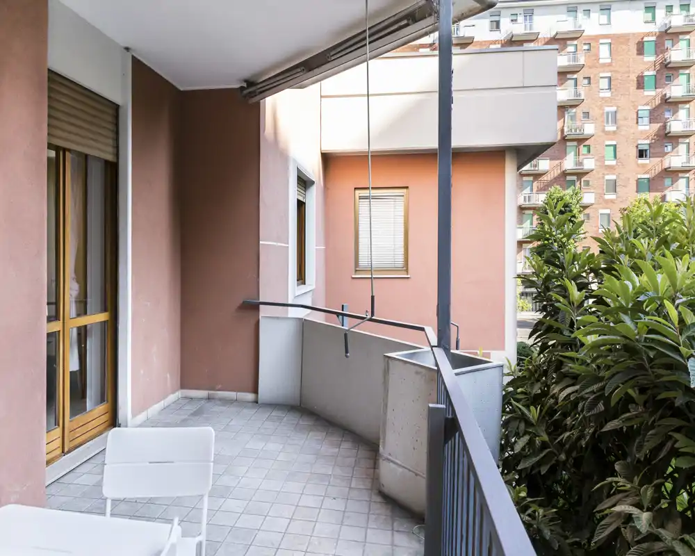 Sunny balcony with tiled floor, metal railing and partial views of neighbouring buildings; small outdoor seating visible and greenery at the edge.