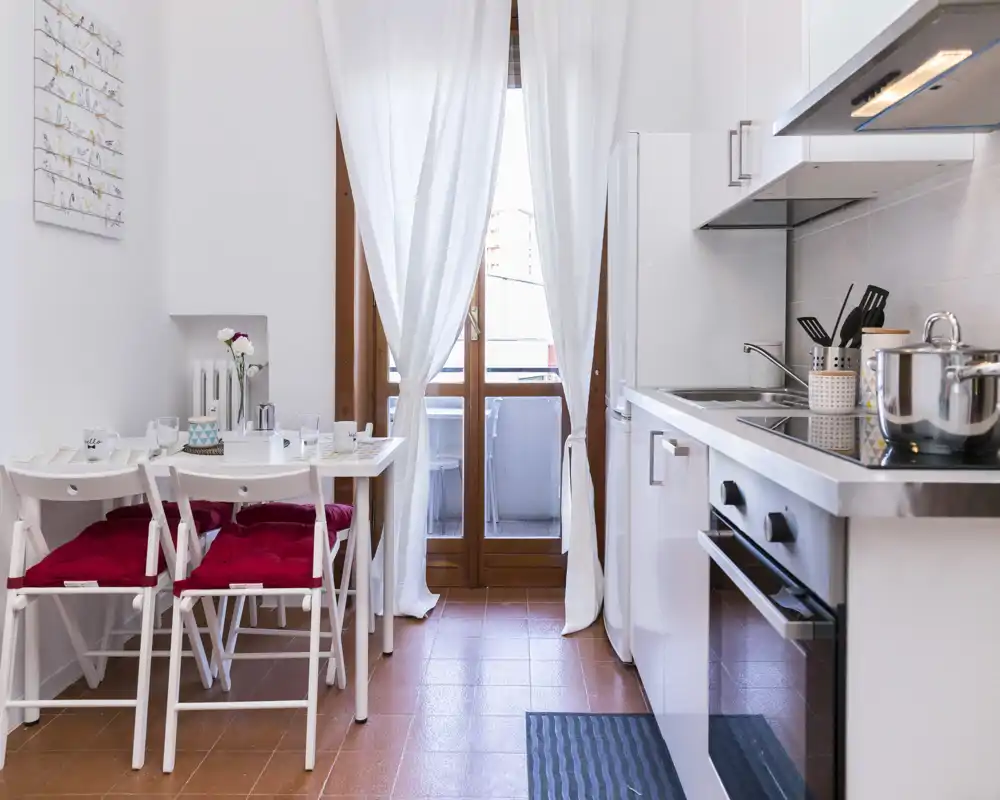 Compact modern kitchen with white cabinets, an electric oven and a small dining table by a bright balcony door — stylish and functional for short stays.