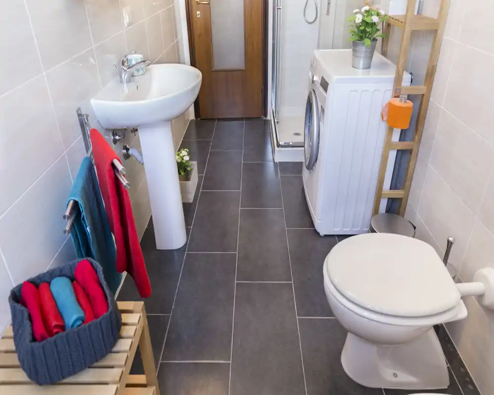 Clean, modern bathroom with a pedestal sink, toilet and compact shower; a washing machine and wooden shelving maximize the small space.