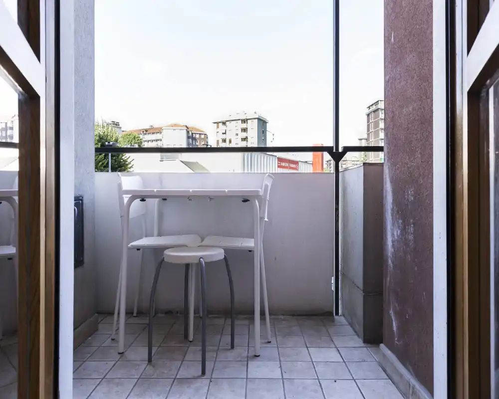 Small private balcony with a tiled floor, a narrow railing-height table and two stools — bright urban view ideal for morning coffee.