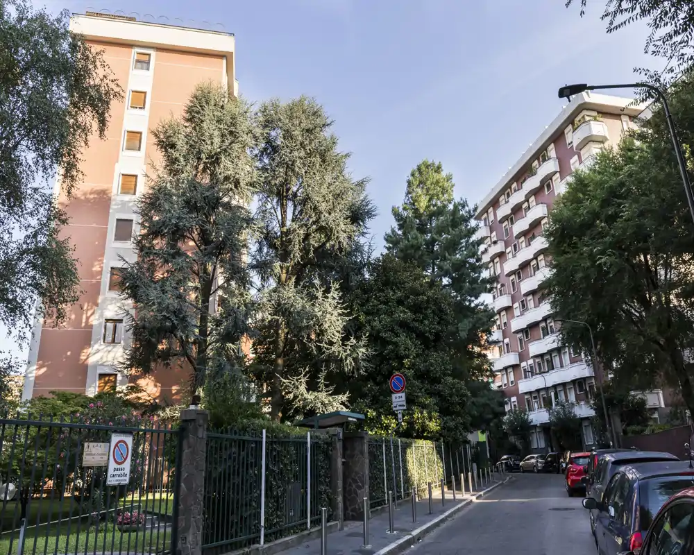 Street-level exterior showing apartment buildings, tall trees and a gated front garden along a calm urban road.