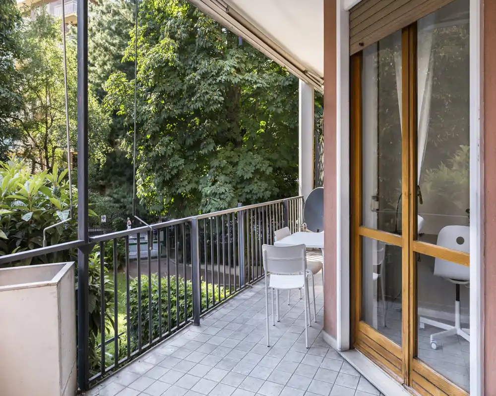 Sunny balcony with tiled floor, metal railing and a small outdoor table with chairs, overlooking leafy trees — ideal for morning coffee.