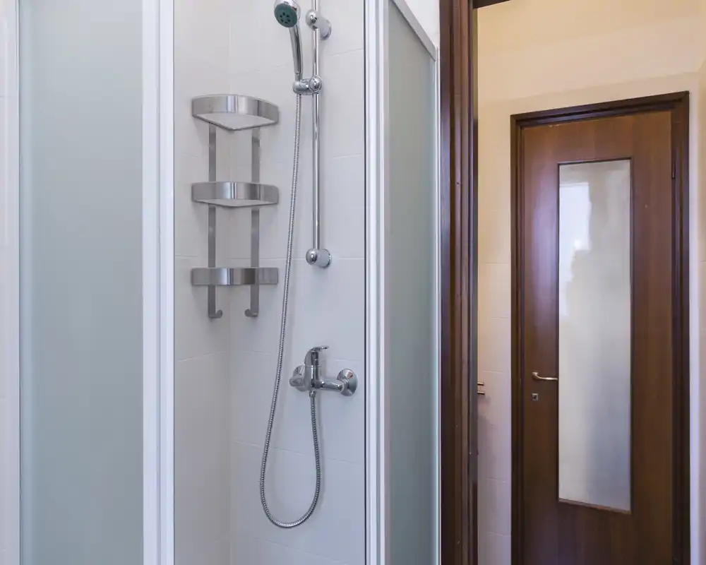 Compact, modern bathroom with a corner shower cubicle, chrome fixtures and a wooden door — clean and well lit.