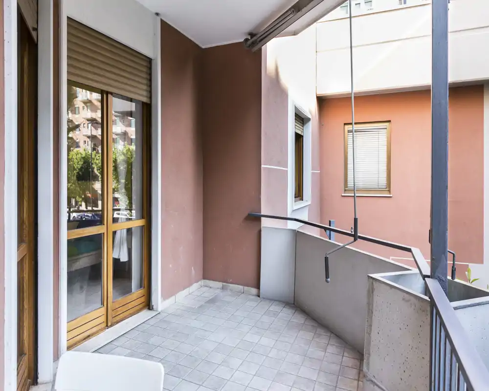 Sunny tiled balcony with protective railing and sliding wooden-framed door leading inside. Suitable for small outdoor seating or drying clothes, with views of neighbouring buildings.