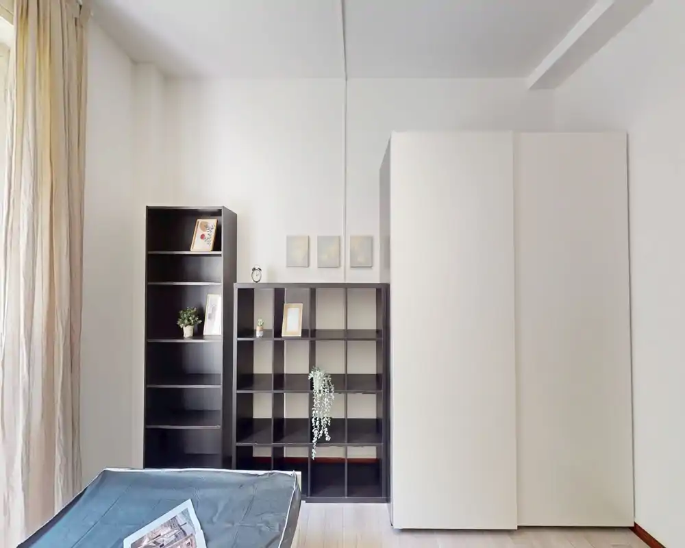 Camera da letto minimalista con un letto basso in primo piano, un armadio bianco alto e scaffalature scure aperte contro una parete luminosa; luce naturale morbida proviene da sinistra.