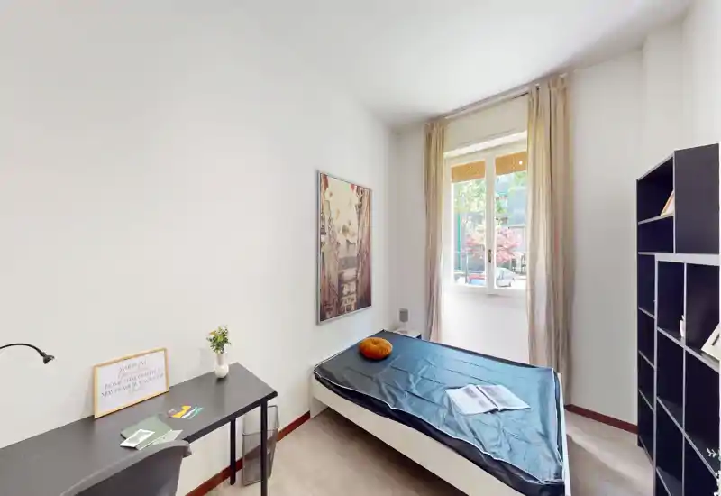Camera singola luminosa e ordinata con finestra, letto compatto, scrivania e scaffalatura—ideale per uno studente o un occupante singolo.