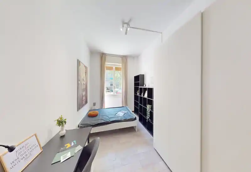 Camera da letto luminosa e compatta con letto singolo vicino alla finestra, scrivania, libreria e armadio — stile moderno e luce naturale.