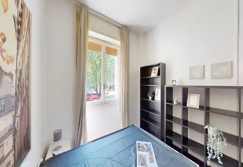 Camera da letto luminosa e ordinata con letto matrimoniale vicino a una grande finestra e scaffalature organizzate, luce naturale gradevole e arredo moderno essenziale.