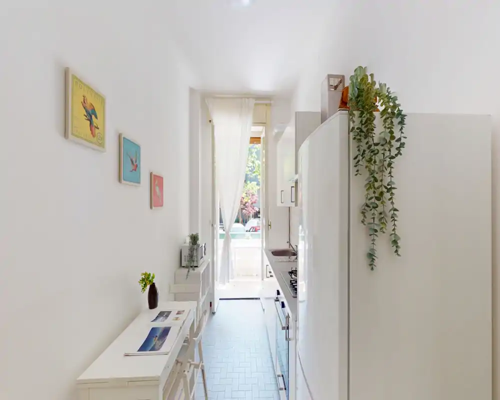 Cucina luminosa e stretta con mobili bianchi, piccolo bancone per la colazione e luce naturale da una porta/terrazzino, decorata con quadretti e una pianta pendente.
