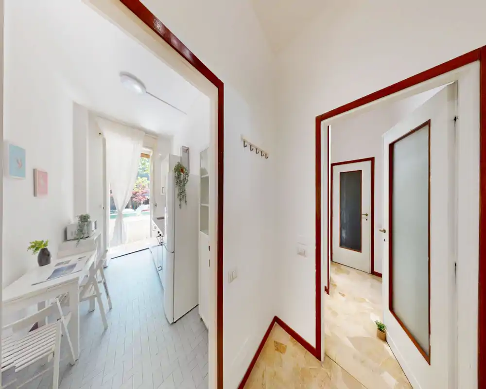 Cucina compatta e luminosa con piccolo tavolo da pranzo, mobili bianchi e una grande finestra/porta che illumina l’ambiente. Stile pulito e minimale con piante e tessili chiari che aggiungono calore.