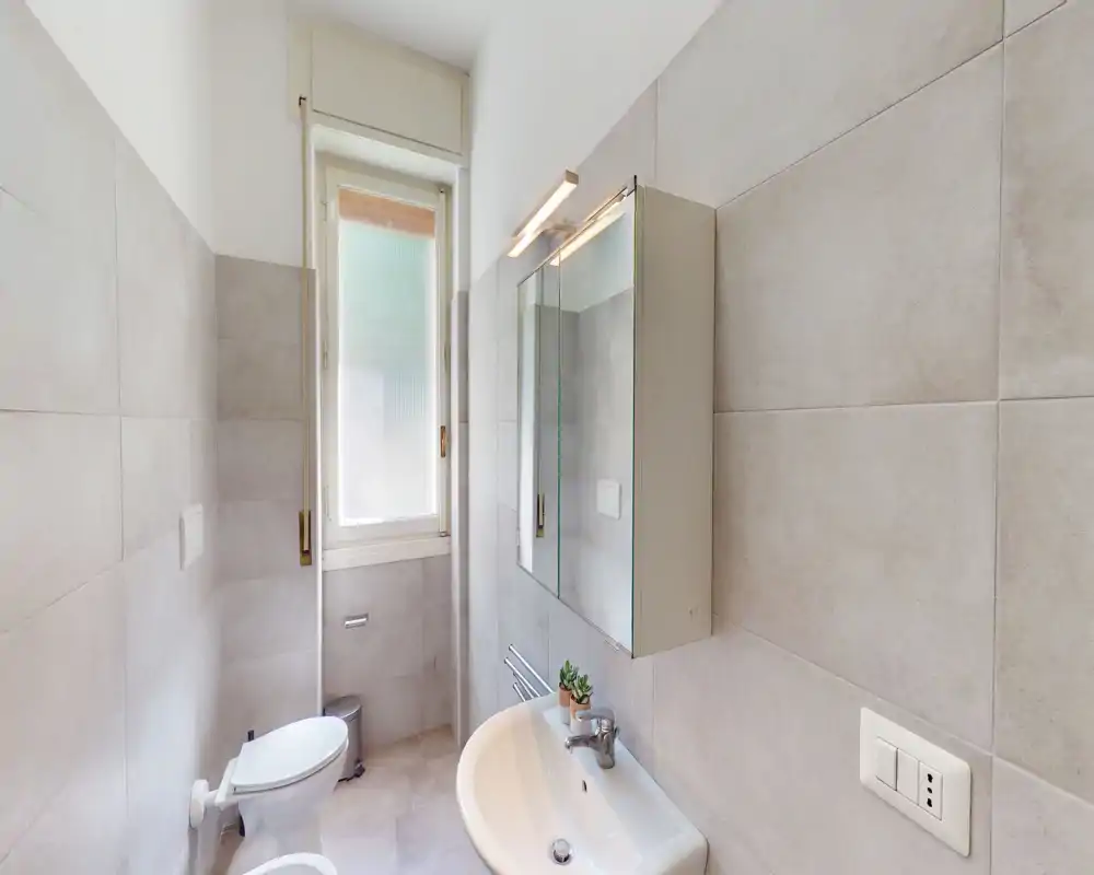 Bagno moderno e luminoso con mobile specchio a parete, lavabo e WC vicino a una finestra alta satinata; piastrelle grandi neutre e sanitari puliti.