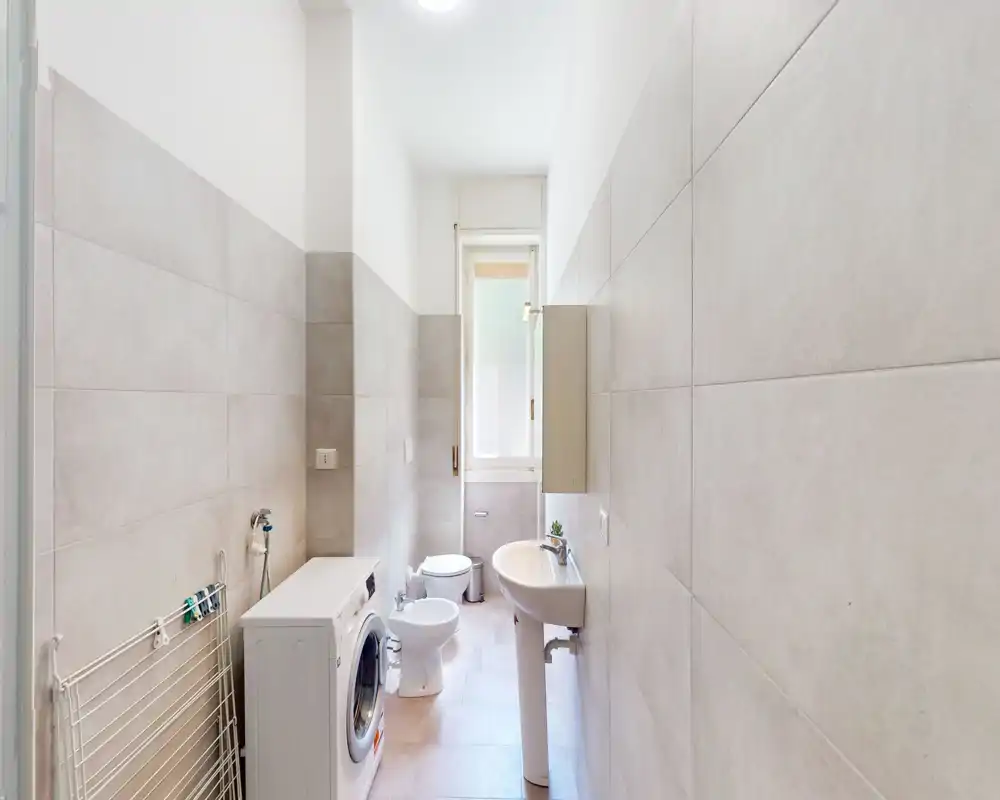 Bagno luminoso e moderno con lavabo a colonna, bidet, WC e lavatrice, illuminato da luce naturale proveniente da una finestra alta.