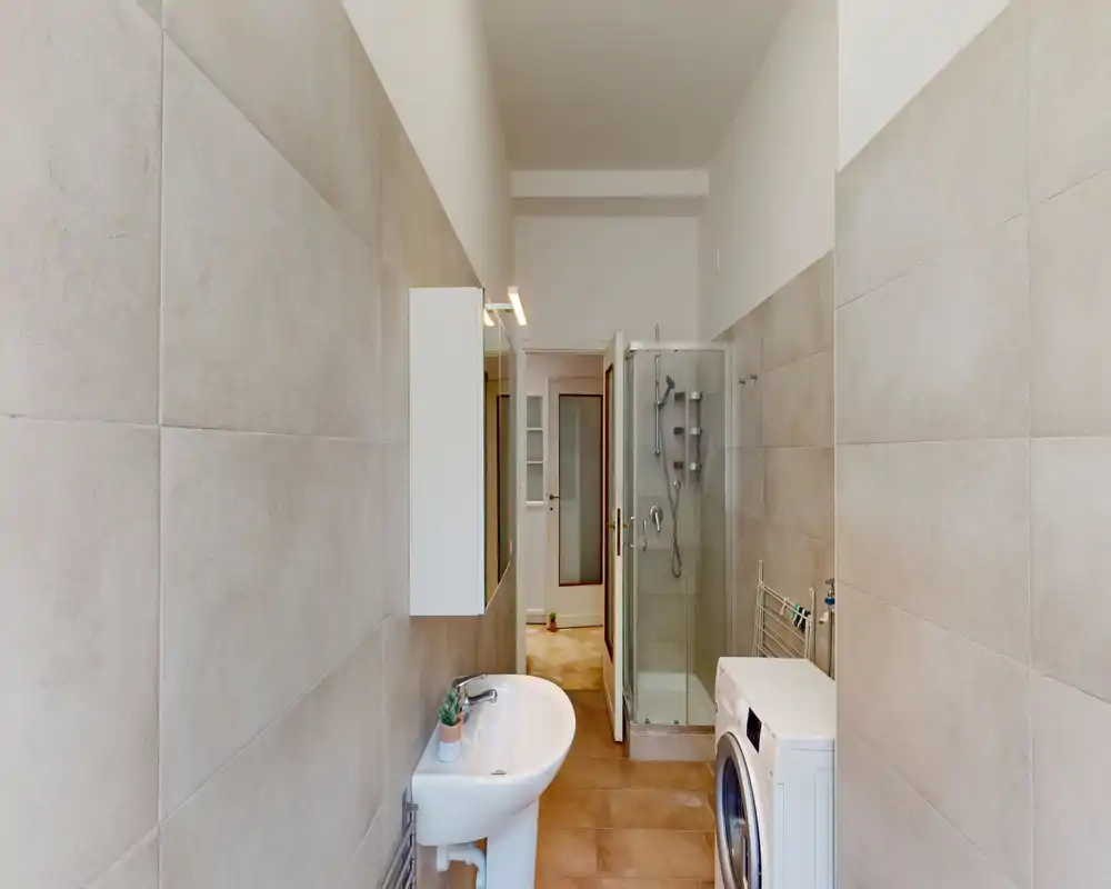 Bagno moderno con cabina doccia in vetro, lavabo a colonna e lavatrice; piastrelle di grande formato dai toni neutri e pavimento caldo per un ambiente pulito e funzionale.