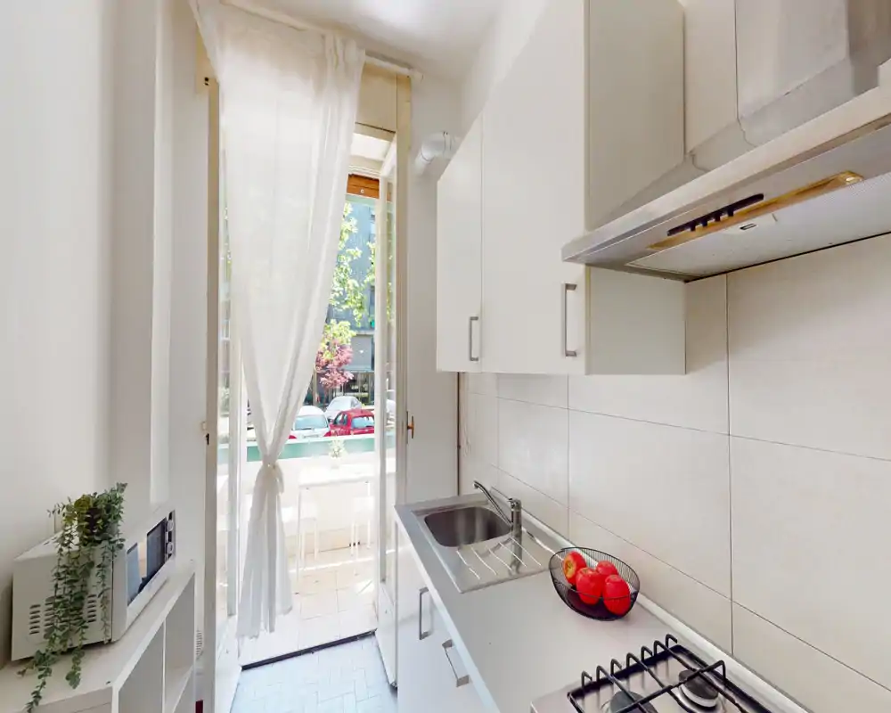 Cucina moderna compatta con mobili bianchi, lavello in acciaio, piano cottura a gas e porta sul balcone che illumina l'ambiente; arredata con una ciotola di frutta e una piccola pianta.