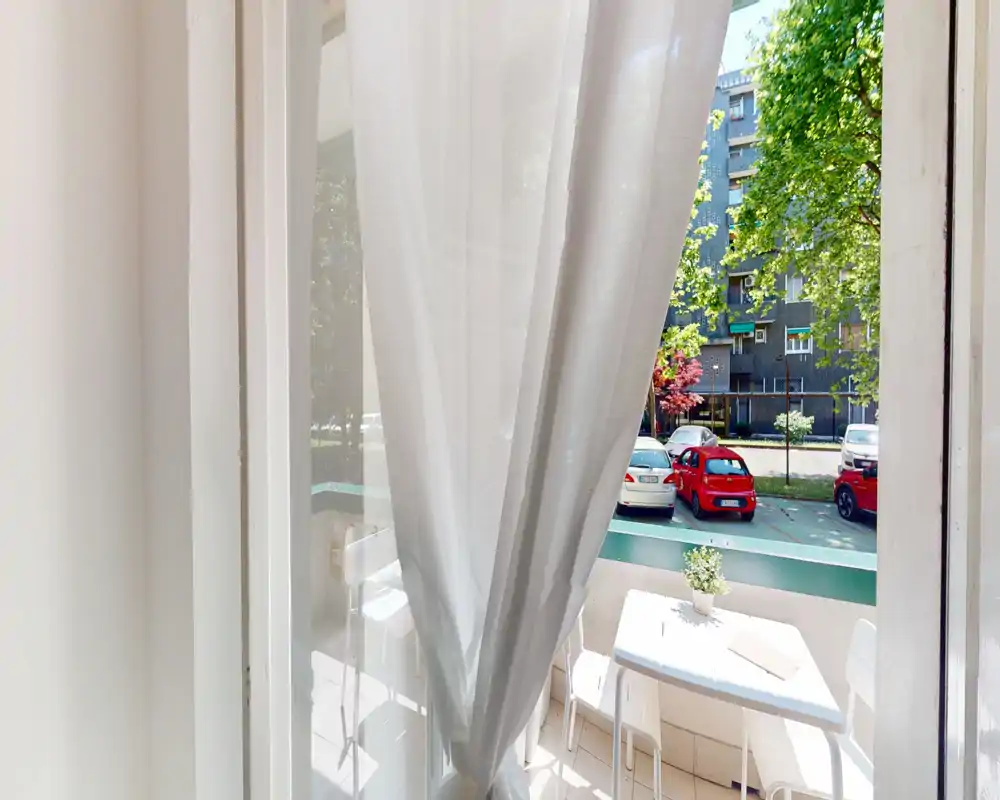 Piccolo balcone soleggiato con tavolo, sedie e tenda di voile che si affaccia su una strada alberata — perfetto per il caffè mattutino.