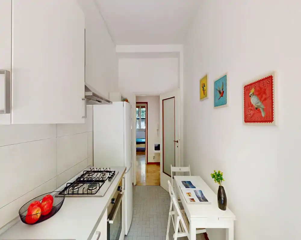 Cucina a corridoio moderna e compatta con mobili bianchi, piano cottura a gas, frigorifero e piccolo tavolo da pranzo; luminosa e ben curata.