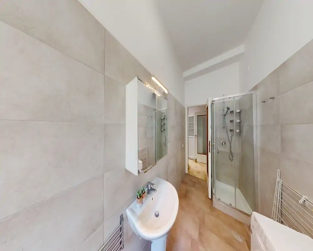 Bagno moderno e ben illuminato con lavabo a colonna, mobile specchiato, doccia con box in vetro e grandi piastrelle dal tono neutro.