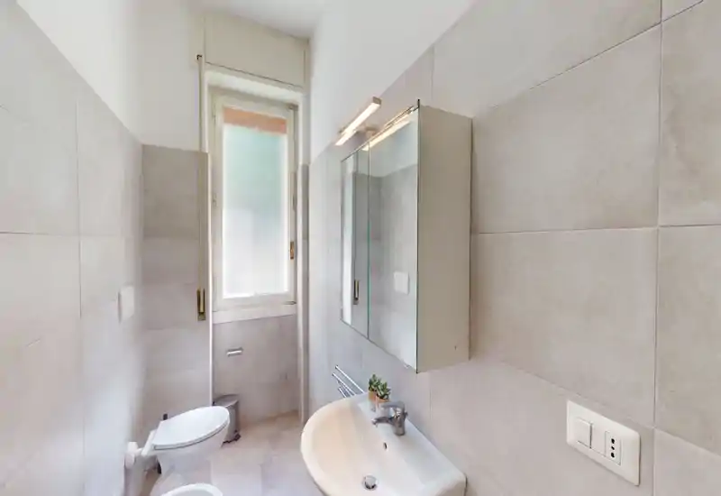 Bright, modern bathroom with wall-mounted mirror cabinet, sink and toilet next to a tall frosted window; neutral large-format tiles and clean fixtures.