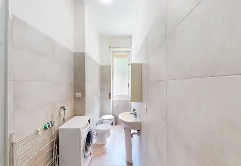 Bright, modern bathroom with pedestal sink, bidet, toilet and washing machine under high natural light from a tall window.