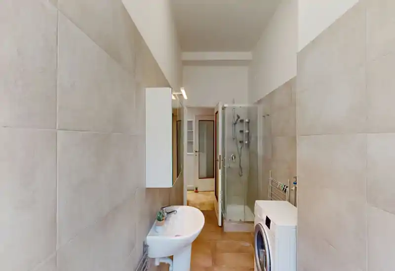 Modern bathroom with a glass shower cabin, pedestal sink and a washing machine; neutral large-format tiles and warm floor tiles create a clean, functional space.