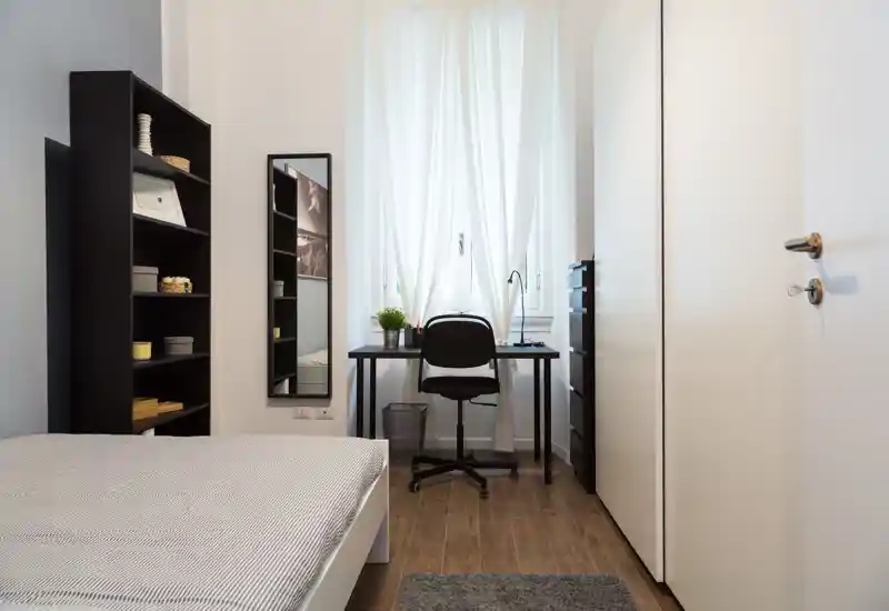Dormitorio moderno y luminoso con la cama en primer plano, un escritorio compacto bajo la ventana y muebles altos para mantenerlo ordenado.