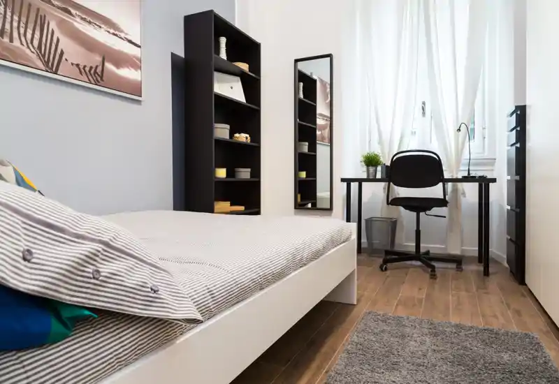 Dormitorio luminoso y ordenado con cama moderna, estantería y un pequeño escritorio junto a la ventana — ideal para descansar y teletrabajar.