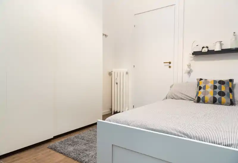 Dormitorio luminoso y minimalista con cama individual bien hecha, cojín geométrico y armario blanco: ideal para alquileres compactos.