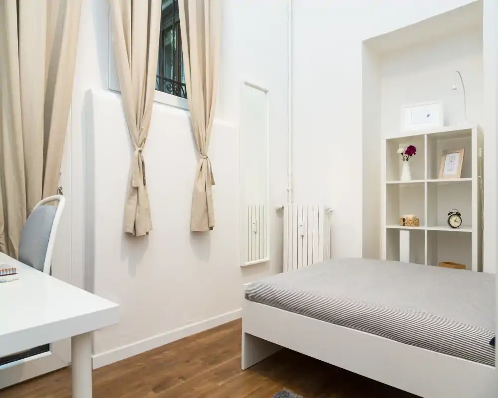 Camera da letto luminosa e ordinata con letto basso, scaffalatura e una piccola scrivania. Toni neutri e luce naturale creano un ambiente calmo e minimal ideale per affitti brevi.