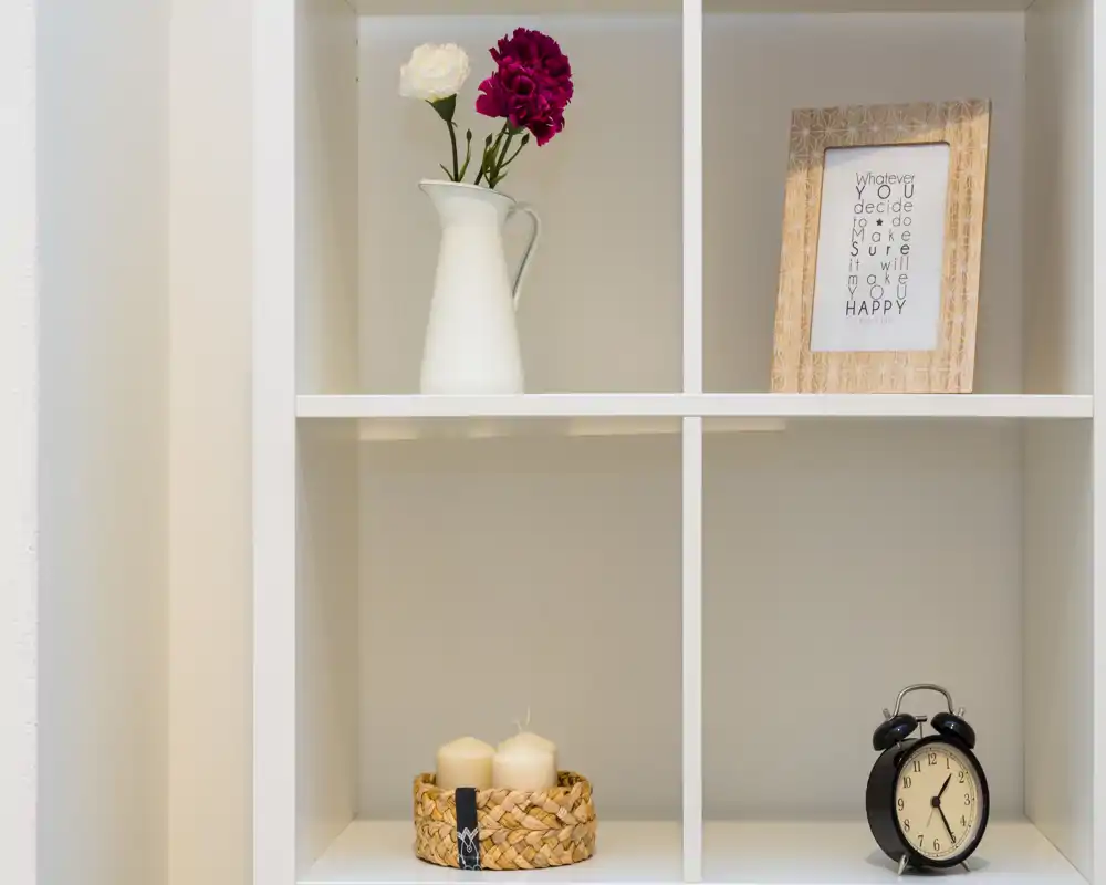 Primo piano di una libreria a cubi bianca con oggetti decorativi: una brocca bianca con fiori, una cornice con stampa, un piccolo cestino con candele e una sveglia vintage.