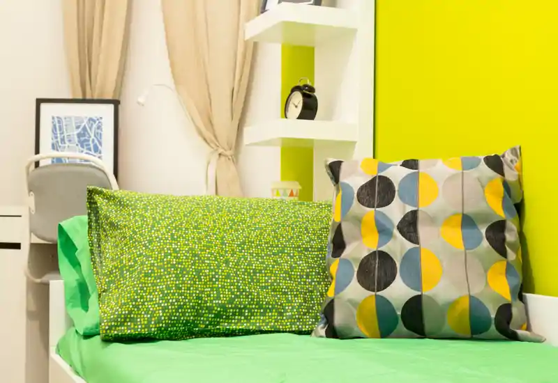 Bright, modern single bedroom corner with colorful pillows and a neatly made bed against a vivid lime wall — ideal for a cozy short-stay.
