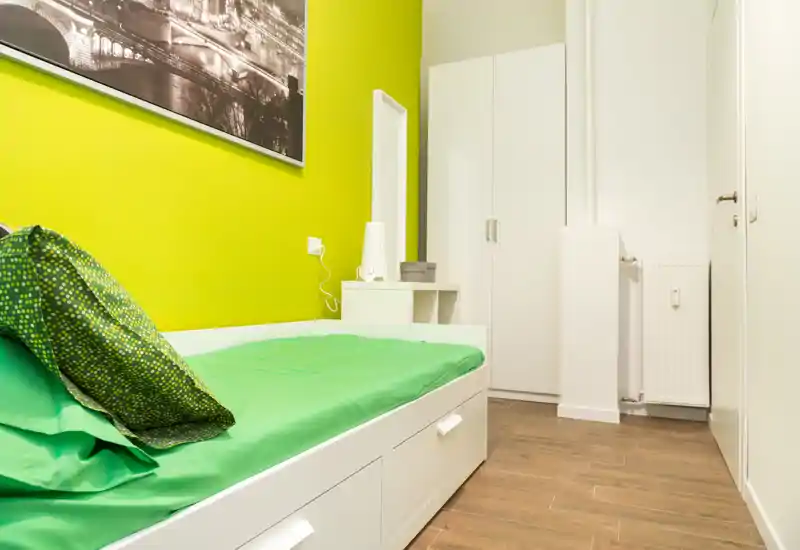 Compact modern single bedroom with a white bed frame, green bedding and a wardrobe. Bright, clean styling with wood flooring and simple decorative accents.