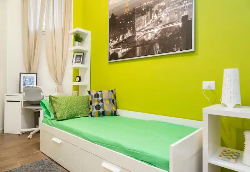 Bright, modern single bedroom with a white daybed, colorful cushions and a clean workspace by the window. The fresh green accent wall and coordinated textiles create a lively, welcoming feel ideal for short-term rentals.