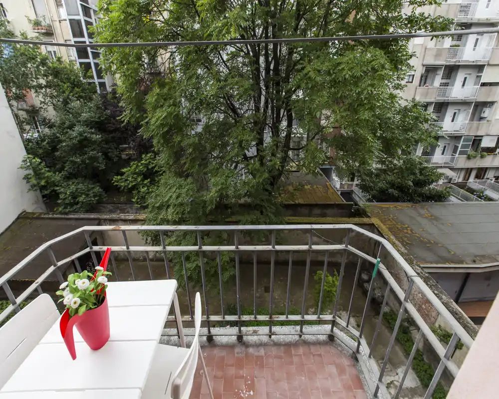 Petit balcon privé avec sol carrelé, rambarde métallique et une table blanche avec un pot rouge décoratif ; donnant sur une cour arborée.