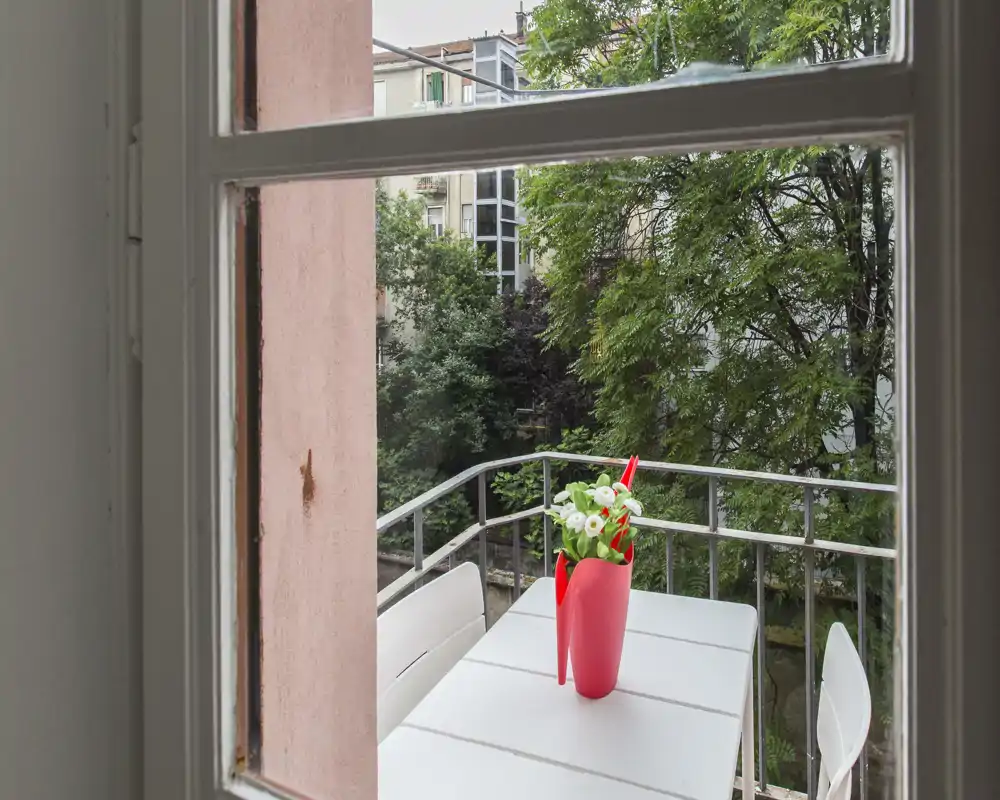 Petit balcon meublé vu depuis une fenêtre, avec une table blanche, deux chaises et un vase rouge fleuri donnant sur des arbres et des immeubles voisins.