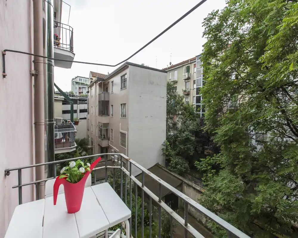 Petit balcon en ville avec une table pliante blanche et un arrosoir rouge décoratif; vue sur les immeubles voisins et une cour verdoyante.