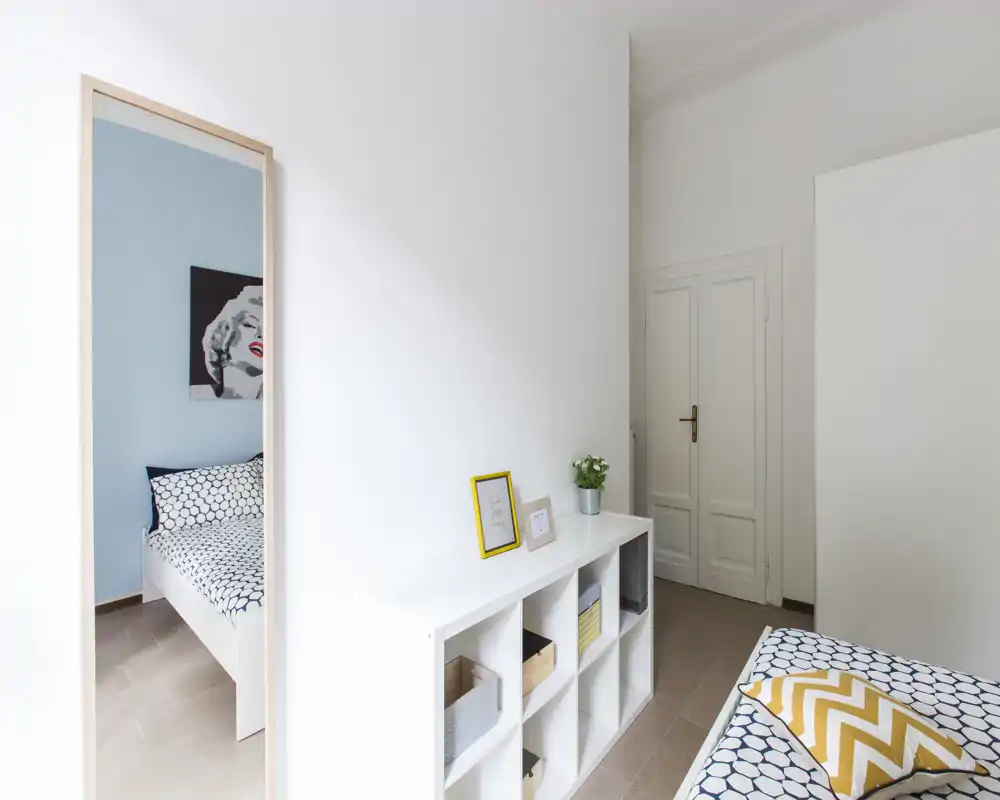 Chambre moderne et lumineuse avec linge de lit à motifs et coussin jaune décoratif; étagères blanches et miroir plein pied reflètent l'espace pour plus de profondeur.