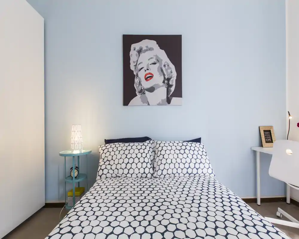 Chambre lumineuse et moderne avec lit double à motifs centré sous une œuvre murale ; lampe de chevet et petit bureau offrent un espace confortable et fonctionnel.