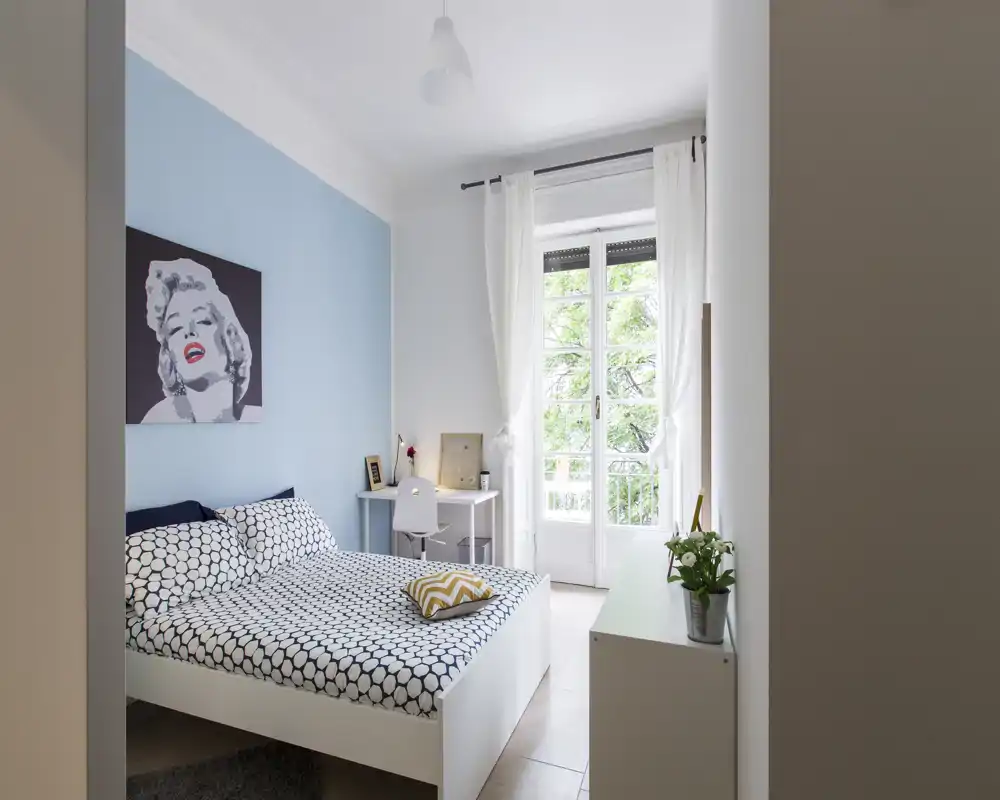 Chambre moderne et lumineuse avec lit double à motif, textiles confortables et un bureau près d'une grande fenêtre donnant sur la verdure.