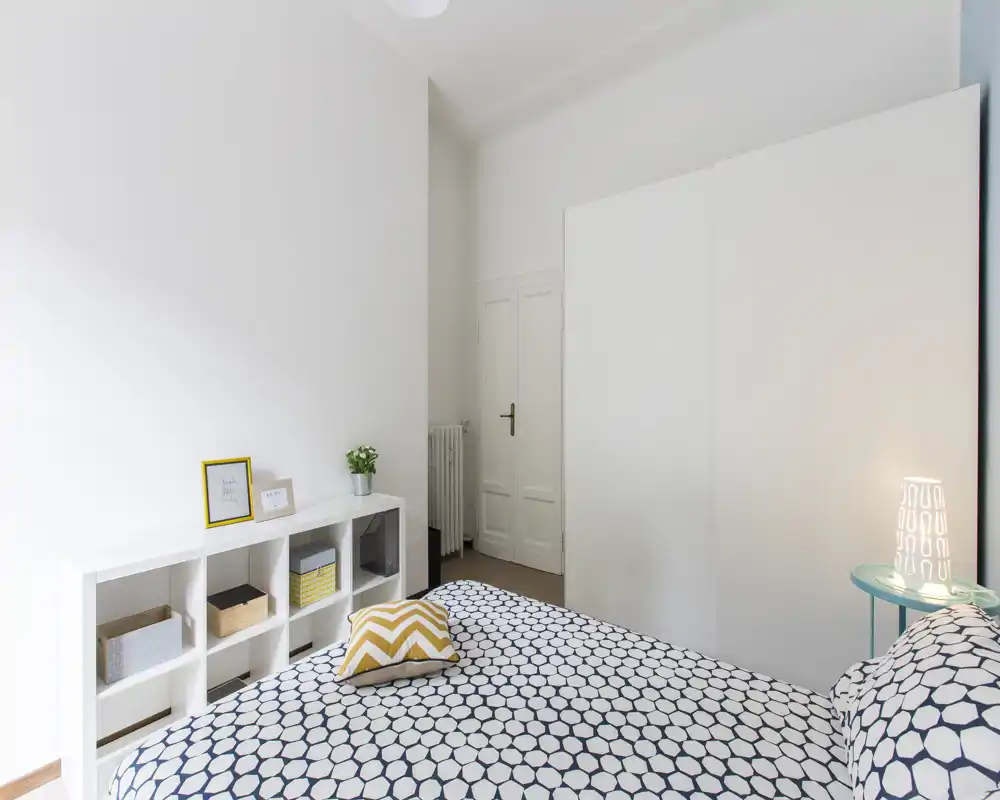 Chambre lumineuse et minimaliste avec un lit double à motif, une grande armoire blanche et des étagères simples ; touches cosy avec un coussin décoratif, une lampe de chevet et une petite plante.