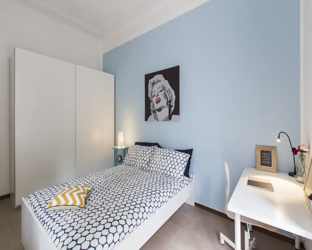 Chambre moderne et lumineuse avec lit double habillé d'une couette à motifs, grand placard blanc, bureau et lumière douce — bien mise en scène pour une annonce.