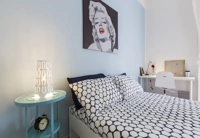 Dormitorio moderno y luminoso con ropa de cama estampada, lámpara de mesita y pequeño escritorio; un cuadro sobre la cama aporta estilo y calidez.