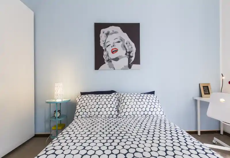 Dormitorio luminoso y moderno con cama doble estampada centrada bajo un cuadro; lámpara de noche y pequeño escritorio crean un espacio acogedor y funcional.