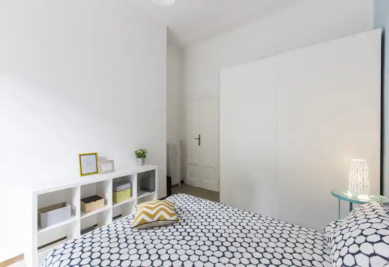 Dormitorio luminoso y minimalista con cama doble estampada, gran armario blanco y estantería sencilla; detalles acogedores como cojín decorativo, lámpara de mesita y una pequeña planta.