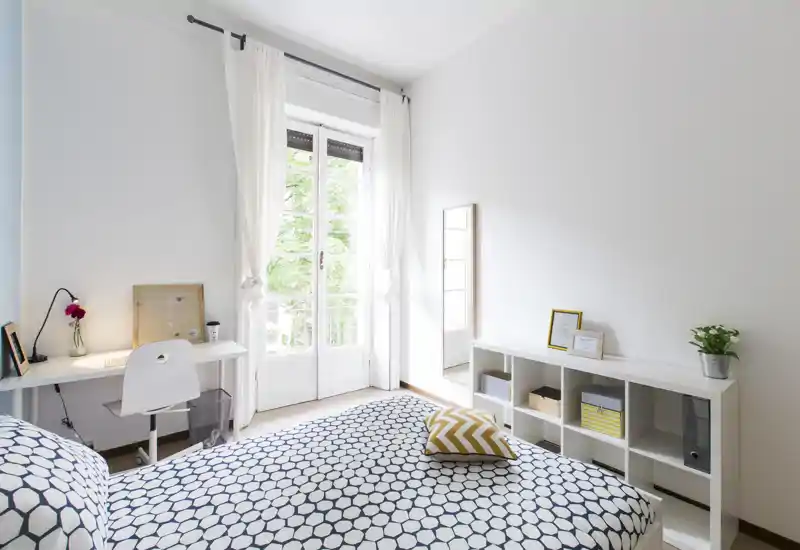 Dormitorio luminoso y minimalista con colcha estampada, escritorio junto a la ventana y estantería modular — mucha luz natural desde las puertas-balconeras y decoración moderna.